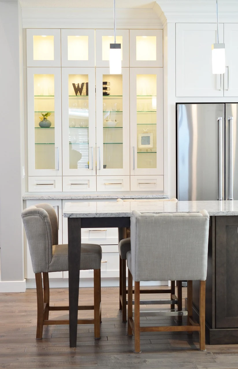 White kitchen cabinetry, a stone counter and upholstered stools beside a stainless refrigerator